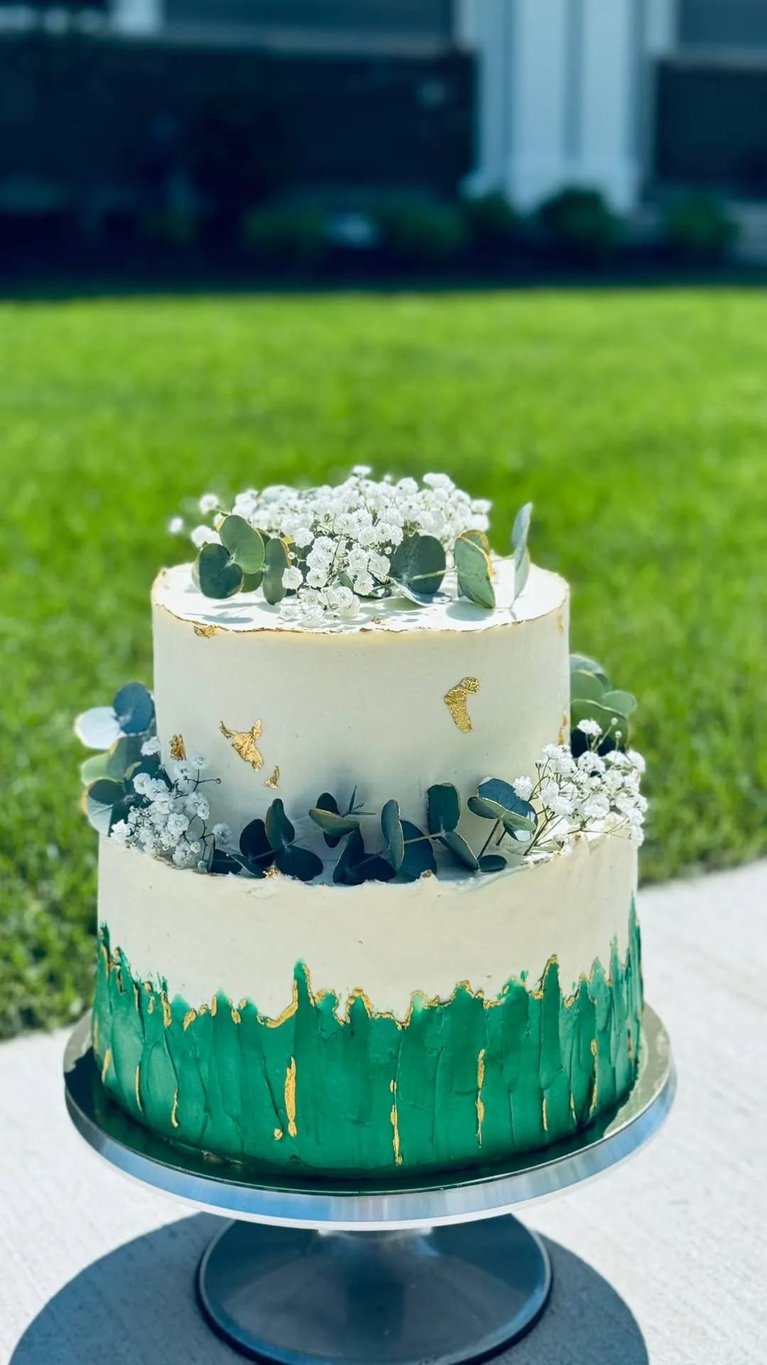 Two-tier wedding cake with eucalyptus, baby's breath, and gold leaf on lush green grass